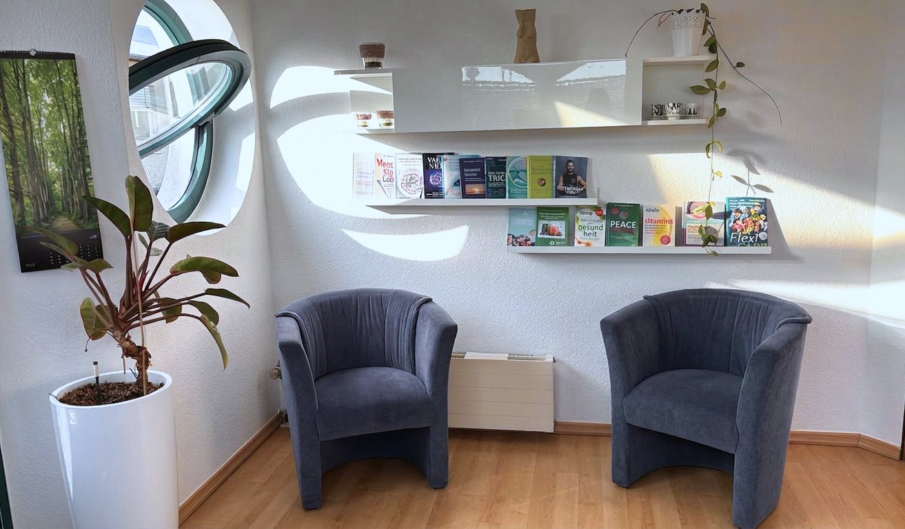 Wartezimmer: zwei Sessel unter Bücherregalen, links ein rundes Fenster und eine große Grünpflanze